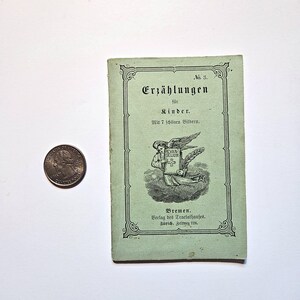 以下が含まれることがあります： 「Erzählungen für Kinder」というタイトルと「Mit 7 schönen Bildern.」というテキストが書かれた緑色の本の表紙。本の表紙には、本を手にした天使の白黒のイラストが描かれており、本には「EVAN-GELIUM」というテキストが書かれている。