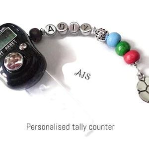 Op de afbeelding: Een zwarte digitale telteller met een doorzichtige plastic band en een kralenketting met de letters "ADIY" en een voetbalbedel. De ketting heeft blauwe, groene en rode kralen. De teller heeft de woorden "Tally Counter" en "Count Reset" erop gedrukt.