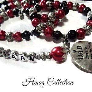 以下が含まれることがあります： 赤と黒のビーズネックレス。シルバーのチャームには「DAD You are someone to look up to no matter how tall I grow」と刻まれています。ネックレスには、シルバーのビーズに「EYAAN」の文字が刻まれています。