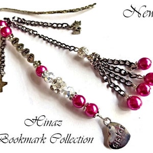 May include: A decorative bookmark with a silver finish, featuring a heart charm that says "sister". The bookmark has pink beads, clear crystals, and dangling chains with star and cat charms. The text "NAME" is spelled out on the bookmark.
