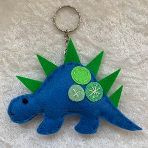 May include: Blue felt dinosaur keychain with green felt spikes and green felt buttons. The dinosaur has a black eye and a silver key ring.