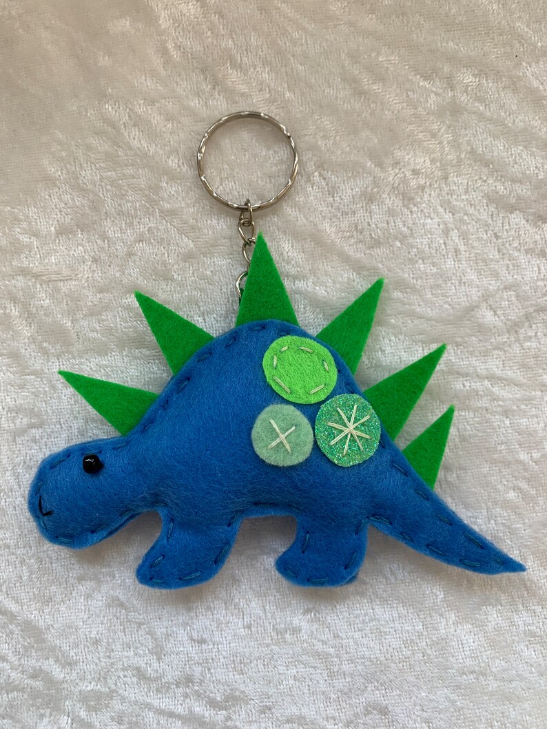 May include: Blue felt dinosaur keychain with green felt spikes and green felt buttons. The dinosaur has a black eye and a silver key ring.