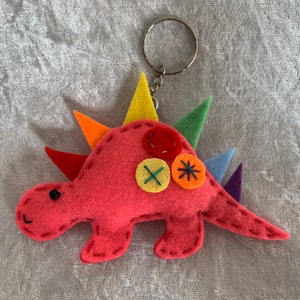 May include: A pink felt dinosaur keychain with rainbow colored spikes and colorful button accents.