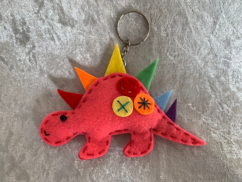 May include: A pink felt dinosaur keychain with rainbow colored spikes and colorful button accents.