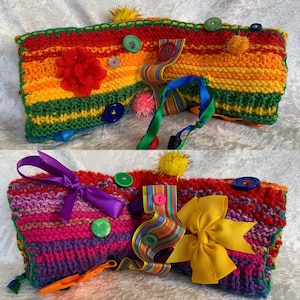 Könnte beinhalten: Zwei bunte gestrickte Babysinnensdecken mit verschiedenen Texturen und Farben. Die obere Decke ist regenbogenfarben gestreift mit grünen, gelben, roten und orangen Streifen. Die untere Decke ist rosa, lila und rot gestreift. Beide Decken haben Knöpfe, Bänder und Bommeln.