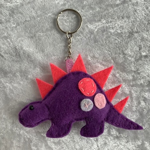 May include: Purple felt dinosaur keychain with pink and white felt accents. The dinosaur has a spiky back and a silver key ring.