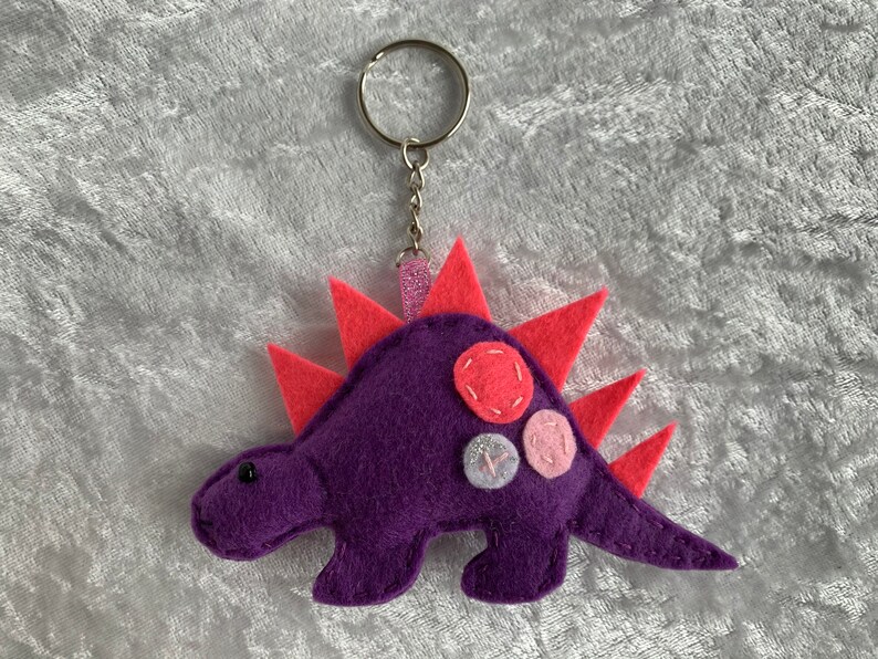 May include: Purple felt dinosaur keychain with pink and white felt accents. The dinosaur has a spiky back and a silver key ring.