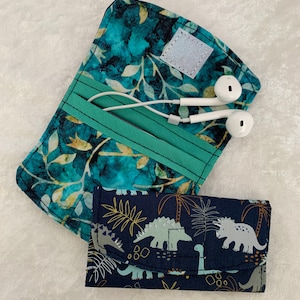 Peut inclure: Deux pochettes en tissu, l'une avec un motif floral bleu sarcelle et or et l'autre avec un fond bleu marine et un imprimé de dinosaure blanc. La pochette sarcelle a une sangle verte et une fermeture velcro blanche. La pochette marine a une fermeture velcro blanche.