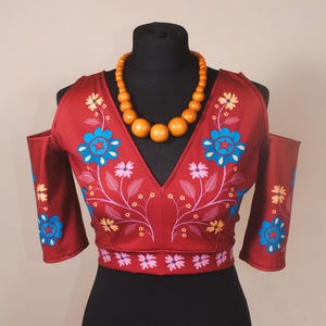 May include: A burgundy off-the-shoulder top with floral embroidery in blue, pink, and yellow. The top has a V-neck and long sleeves. A wooden bead necklace is around the neck.
