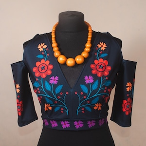 Puede incluir: Top azul oscuro con hombros descubiertos y diseño floral en rojo, naranja, morado y azul. El top se combina con un collar de cuentas de madera. El estampado floral se repite en las mangas y la cintura. El top se exhibe en un maniquí negro.