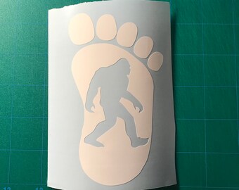 May include: A white vinyl decal featuring a large footprint shape with a silhouette of a Bigfoot creature cut out from the center. The decal is presented on a white backing sheet, showcasing the detailed outline of the toes and the creature figure.