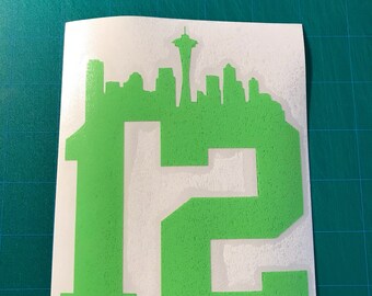 May include: Green vinyl decal featuring the Seattle skyline and the number 12. The skyline includes the Space Needle. The decal is on a white background, with a green cutting mat in the background.
