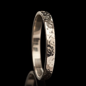 Puede incluir: Un anillo plateado con una superficie texturizada. El anillo tiene un diseño martillado, creando un patrón único. El anillo se encuentra sobre un fondo negro.