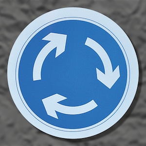 Puede incluir: Señal de tráfico circular azul con borde blanco y tres flechas blancas que indican una rotonda. Las flechas están curvadas y apuntan en el sentido de las agujas del reloj. La señal está sobre un fondo gris.