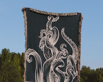 May include: A woven tapestry throw blanket with a dark blue background and a white design of an octopus with tentacles. The blanket is being held up by a person in a grassy field.