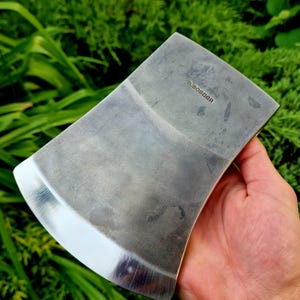 May include: A steel axe head with a polished, curved cutting edge and a matte grey finish on the body. The metal surface features the stamped text D. MOSIER. The axe head is shown from a side profile, highlighting the metallic texture and edge.