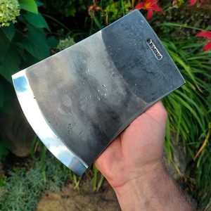 May include: A large, rectangular metal axe head with a polished, curved blade. The axe head has a dark, textured surface and a small, stamped inscription. The axe is held against a backdrop of green foliage and red flowers.