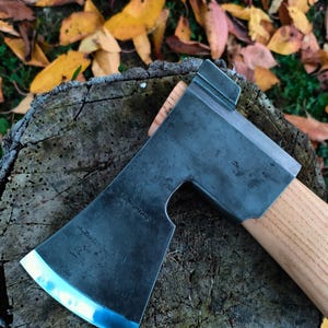 Könnte beinhalten: Eine Stahlaxt mit Holzgriff, die auf einem Baumstumpf liegt. Der Axtkopf ist dunkelgrau mit einer polierten silbernen Kante. Der Griff ist hellbraun. Die Axt ist angewinkelt, die Schneide zeigt zum Betrachter. Der Hintergrund umfasst Herbstlaub.