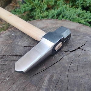 Puede incluir: Un martillo de acero y madera con una cabeza plana y biselada y un mango de madera. El martillo está apoyado sobre una superficie de madera desgastada, con follaje verde al fondo.