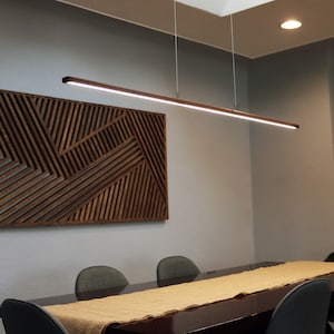 May include: A modern dining room with a long wooden table, four chairs, and a linear pendant light fixture hanging above the table. A geometric wooden wall art piece hangs on the wall behind the table.