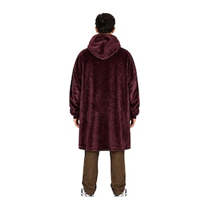 May include: A deep burgundy oversized hooded sweatshirt made of a plush, fuzzy material. The garment features long sleeves with ribbed cuffs, a large hood, and a loose, knee-length fit that hangs straight down, paired with plain brown trousers.