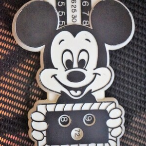 May include: Black and white Mickey Mouse figurine holding a black and white sign with the number 72 and two 8-ball pool balls. The sign has a white border with a black and white striped pattern. The figurine is attached to a white ruler with black numbers.
