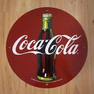 May include: A red circular Coca-Cola sign with a white Coca-Cola logo and a green and brown Coca-Cola bottle. The sign has the text "© 2003 The Coca-Cola Company. All Rights Reserved."