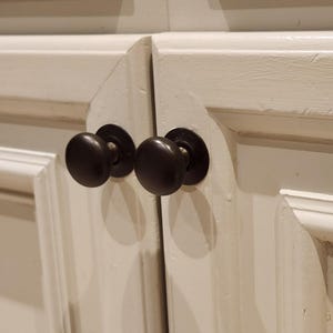 May include: Two black cabinet knobs with a round shape and a slightly raised edge. The knobs are mounted on a white cabinet door.