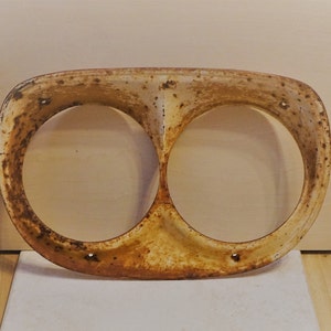 May include: A vintage, rusty, white metal headlight housing with two circular openings. The housing is slightly curved and has a worn, aged appearance.