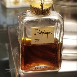 May include: A clear glass perfume bottle containing amber liquid with a label reading Réplique Raphael Paris. The bottle features a gold cord wrapped around the neck and a clear glass stopper with the letter R embossed on the top surface.