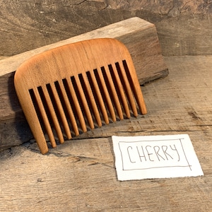 K&ouml;nnte beinhalten: Ein Kamm aus Kirschholz. Der Kamm hat eine abgerundete Form und ist aus hellem braunem Holz gefertigt. Der Kamm liegt auf einer Holzoberfl&auml;che. Der Text "CHERRY" ist auf einem Blatt Papier neben dem Kamm geschrieben.