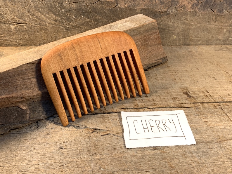 K&ouml;nnte beinhalten: Ein Kamm aus Kirschholz. Der Kamm hat eine abgerundete Form und ist aus hellem braunem Holz gefertigt. Der Kamm liegt auf einer Holzoberfl&auml;che. Der Text "CHERRY" ist auf einem Blatt Papier neben dem Kamm geschrieben.