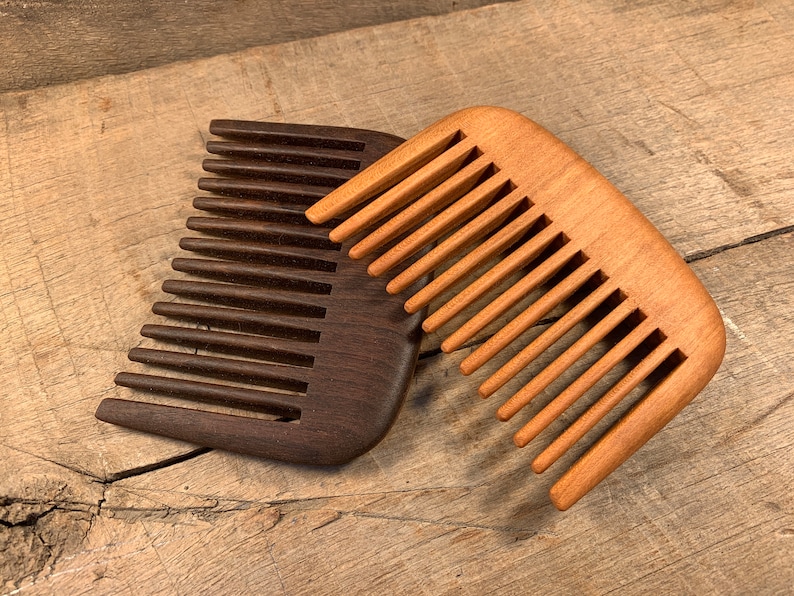 K&ouml;nnte beinhalten: Zwei Holzk&auml;mme, einer dunkelbraun und einer hellbraun, liegen auf einer Holzoberfl&auml;che. Die K&auml;mme haben eine abgerundete Form und sind aus Naturholz gefertigt.