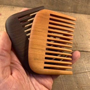 K&ouml;nnte beinhalten: Zwei Holzk&auml;mme, einer braun und einer hellbraun, mit abgerundeten Kanten und feinen Z&auml;hnen. Die K&auml;mme werden in einer Hand gehalten.