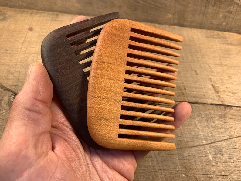K&ouml;nnte beinhalten: Zwei Holzk&auml;mme, einer braun und einer hellbraun, mit abgerundeten Kanten und feinen Z&auml;hnen. Die K&auml;mme werden in einer Hand gehalten.