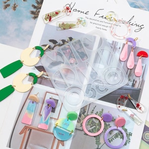 Pode incluir: Um molde de silicone transparente com várias formas para fazer brincos. O molde é rodeado por brincos de resina coloridos com diferentes designs, incluindo formas geométricas, flores e padrões abstratos.