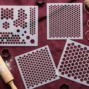 May include: Four white plastic stencils with honeycomb patterns, a rolling pin, and cookie cutters are arranged on a burgundy surface. The stencils feature various honeycomb designs for baking or crafts.