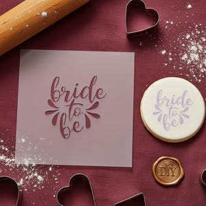 May include: A stencil and a decorated biscuit, both with the words "bride to be", alongside heart-shaped cookie cutters. The items are on a burgundy surface with flour and a rolling pin.