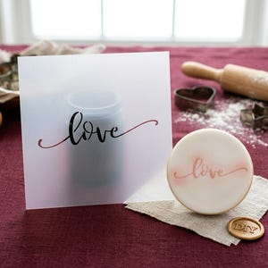 May include: A frosted square card with the word "love" in black script is next to a frosted cookie with the word "love" in pink script. A rolling pin, heart-shaped cookie cutter, and a "DIY" wax seal are also visible.