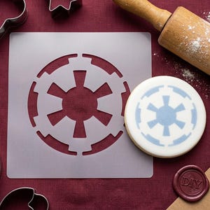 Peut inclure: Un pochoir en plastique gris avec un motif circulaire, à côté d'un biscuit décoré du même motif bleu. Un rouleau à pâtisserie en bois et des emporte-pièces sont également visibles. Le pochoir est pour décorer les biscuits.