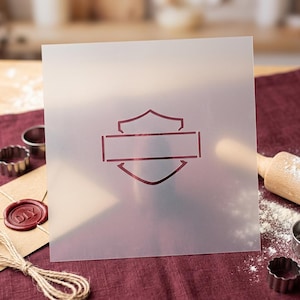 Peut inclure: Un pochoir carré en plastique semi-transparent présentant un motif graphique centré composé d'un écusson traversé par une barre rectangulaire horizontale. Le pochoir est clair avec une finition mate et le motif est découpé en rouge foncé.