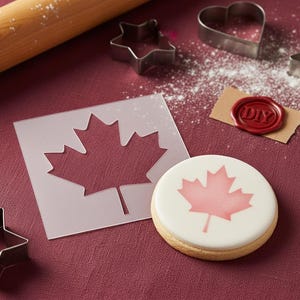 May include: A round biscuit with a red maple leaf design, a stencil, and cookie cutters on a burgundy surface. A rolling pin and a wax seal with "DIY" are also present. The cookie cutters are in the shapes of a star and a heart.