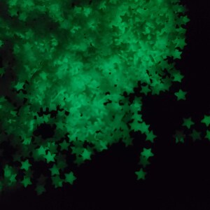 May include: A collection of glowing green star-shaped confetti against a black background. The stars are scattered, creating a sparkling effect. The image is dark, with the stars being the primary source of light.