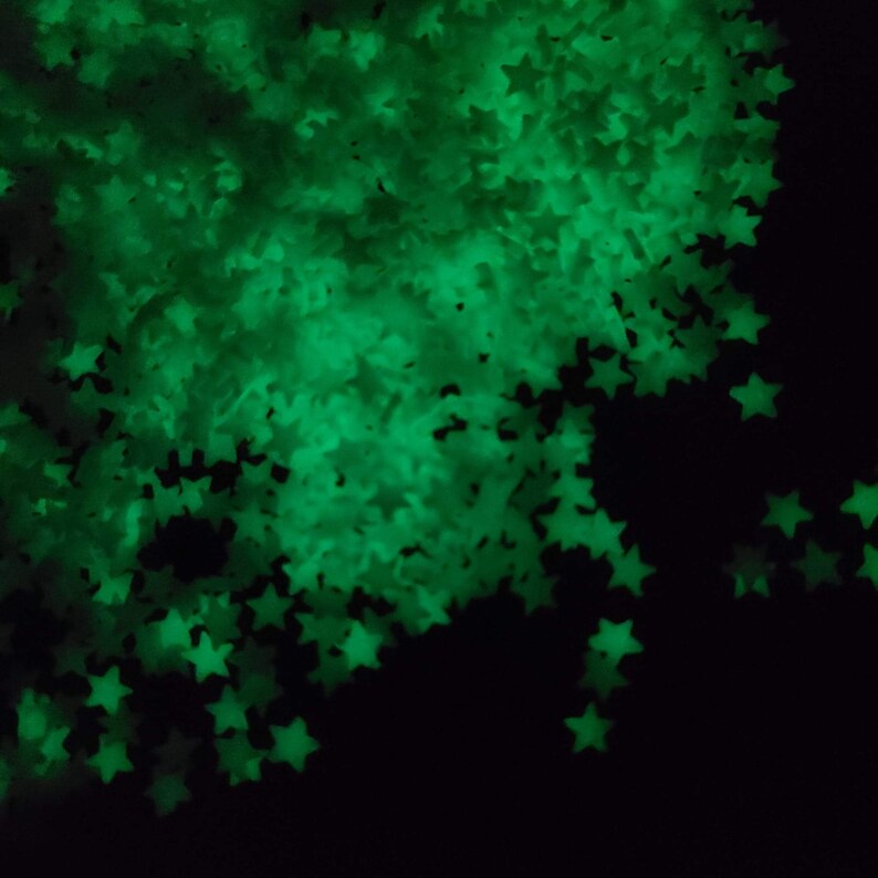 May include: A collection of glowing green star-shaped confetti against a black background. The stars are scattered, creating a sparkling effect. The image is dark, with the stars being the primary source of light.