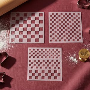 May include: Three square stencils with checkered patterns, plus metal cookie cutters in heart, star, and flower shapes. A rolling pin and a gold-coloured stamp with a whisk design are also present. The items are arranged on a red surface with flour.
