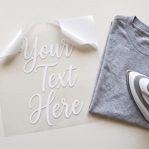 Op de afbeelding: Een grijs t-shirt naast een doorzichtig transfervel met de witte scripttekst Your Text Here, vergezeld van een wit en grijs handstrijkijzer voor warmteoverdracht op het oppervlak van de stof.