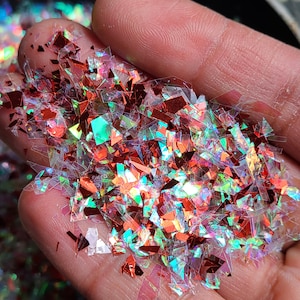 May include: A pile of iridescent red and clear glitter flakes. The glitter is in a variety of shapes and sizes, and it has a holographic finish.