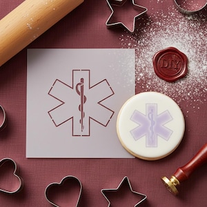 May include: A light purple stencil and a cookie decorated with the Star of Life medical symbol. The stencil is on a light purple background. A rolling pin, cookie cutters, and a DIY wax seal are also present.