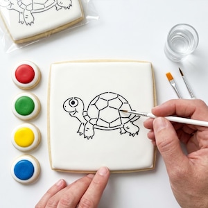 May include: A square biscuit with a black outline of a turtle design is being painted with a paintbrush. The biscuit is on a white surface, with four colourful paint circles and a glass of water nearby. Another biscuit with the same design is in a clear bag.