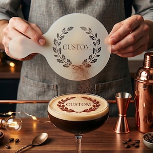 May include: A custom coffee stencil with a laurel wreath design is held over a cocktail. The stencil is used to create a decorative design on the coffee drink. A copper cocktail shaker and other bar tools are in the background.